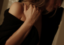 Woman in a black off-the-shoulder sweater holding the back of her neck, face turned away, in dim warm light.